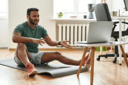 Ein Mann sitzt auf einer Yogamatte und spricht über einen Computer mit jemanden.